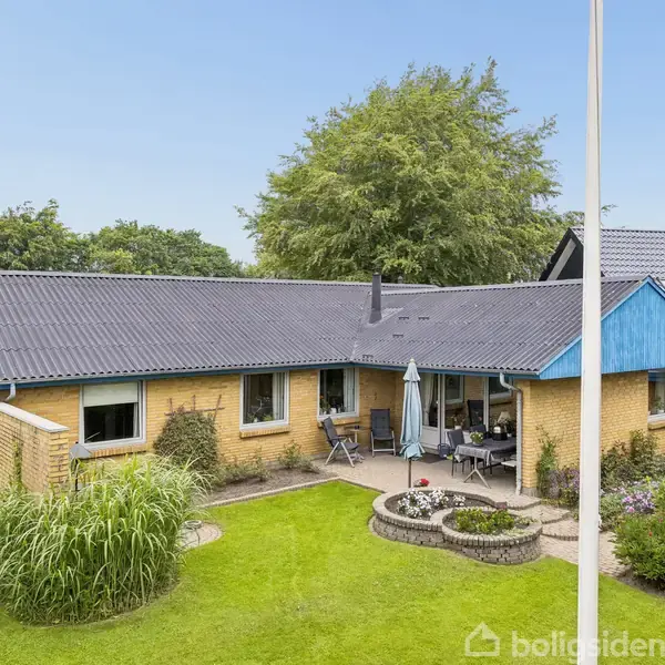 Et gult murstenshus med blå tagdetaljer står i en grøn have. Terrassen er møbleret med bord og parasol. I baggrunden ses store buske og træer.