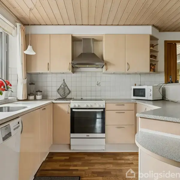 Køkken med træloft og lyse skabslåger, hvor en opvaskemaskine står ved siden af en vask. Ovn, emhætte og mikroovn på køkkenbord med flisevæg og krukkeplanter som dekoration.