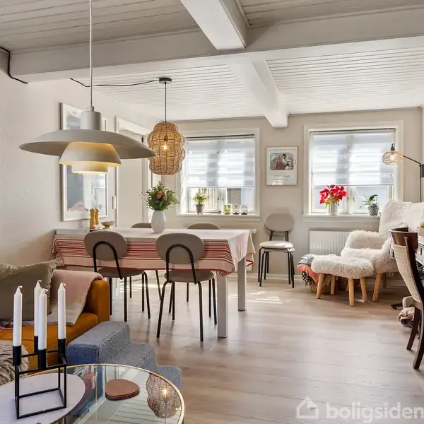 Stue med spisebord dækket af en stribet dug, omgivet af stole. Store vinduer lader lys ind. En gul sofa står til venstre. Dekorative elementer og billeder præger rummet.