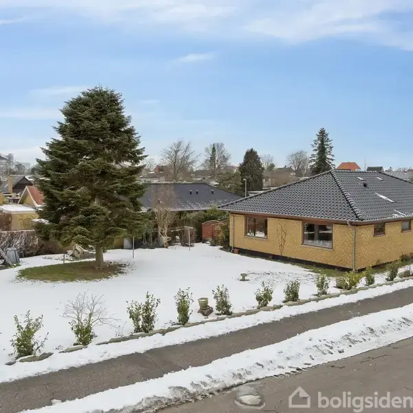 Et murstenshus med sort tag står i en snedækket have. En stor grantræ står til venstre. Omgivelserne er et villakvarter med flere boliger og træer.