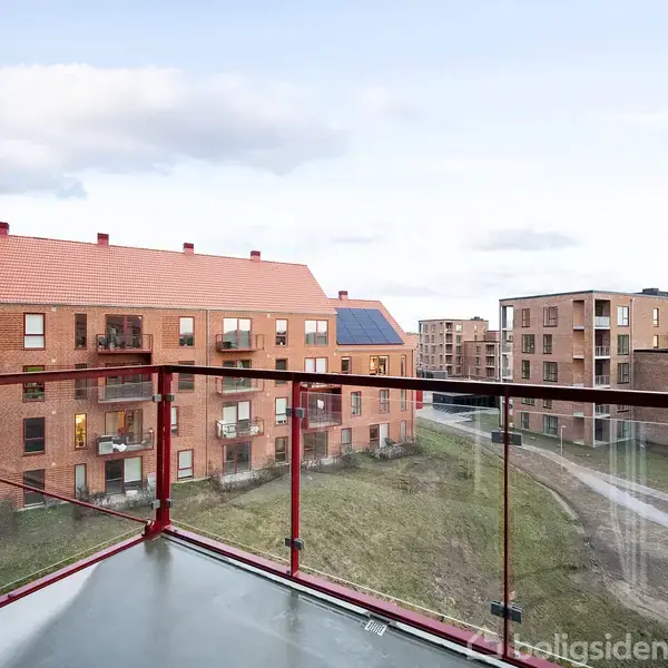 En balkon med glasrækværk viser rødbrune murstensbygninger i en boligkvarter. Bygningerne har store vinduer og balkoner. Stier og græsarealer ses mellem bygningerne under en lys himmel.