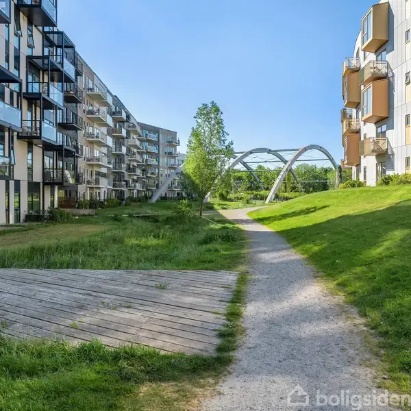 Flere moderne lejlighedsbygninger står ved siden af en grussti, omgivet af grønne græsarealer og træer, med en bueformet metalbro i baggrunden under en klar blå himmel.