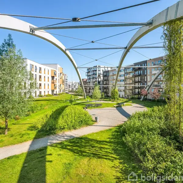Grønne områder ligger mellem moderne lejlighedsbygninger under buede metalstrukturer. Sollys oplyser parken, mens stier krydser plænerne, og træer tilføjer naturlige elementer til urbane omgivelser.