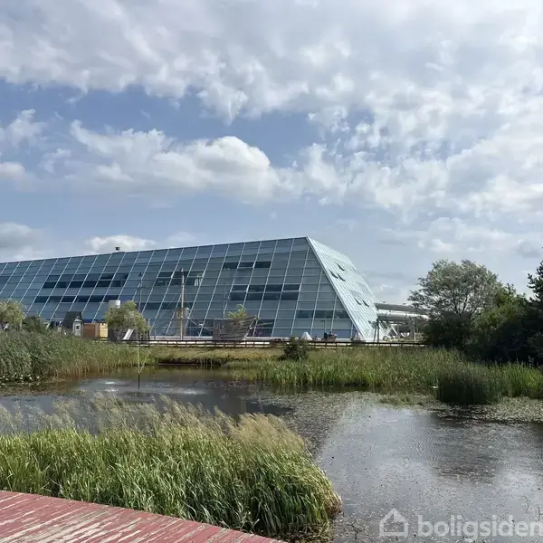 En moderne, skrånende glasbygning reflekterer sollys, omgivet af grøn vegetation og en stille dam under en delvis overskyet himmel.