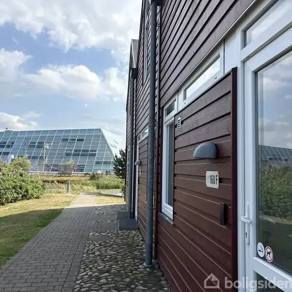 En træbeklædt bygning med en dør mærket "101F" står ved siden af en brostensbelagt sti. I baggrunden ses en stor glasbygning og en græsplæne under en blå himmel.