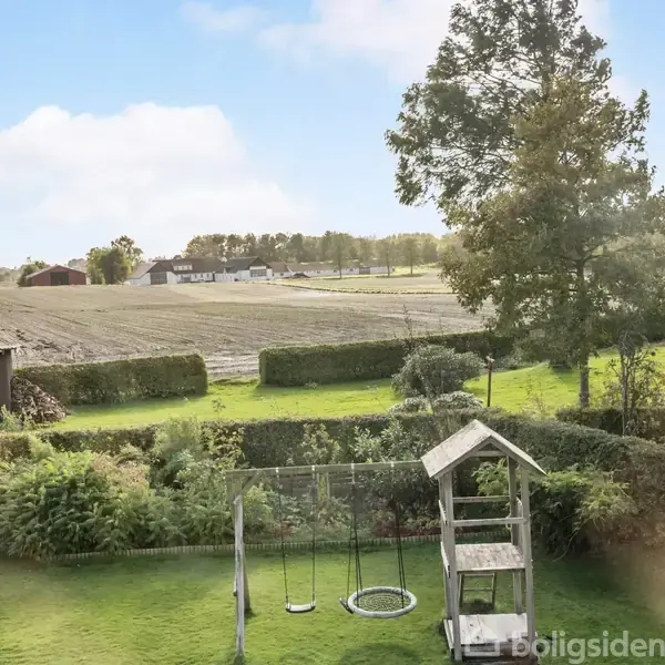 Gyngestativ står stille i en have med grøn græsplæne. Omkring er der buske, træer og åbne marker i baggrunden under en blå, let overskyet himmel.