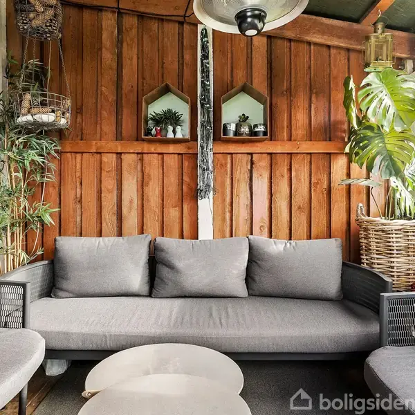 En grå sofa står på en terrasse omgivet af trævægge og planter. To minimalistiske hylder med dekoration hænger på væggen. En stor plante står i en flettet kurv.