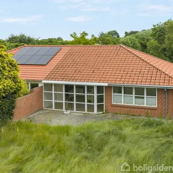Et murstenshus med rødt tegltag og solpaneler på taget står stille. Huset er omgivet af høj græs og træer, hvilket skaber en grøn og naturlig atmosfære.