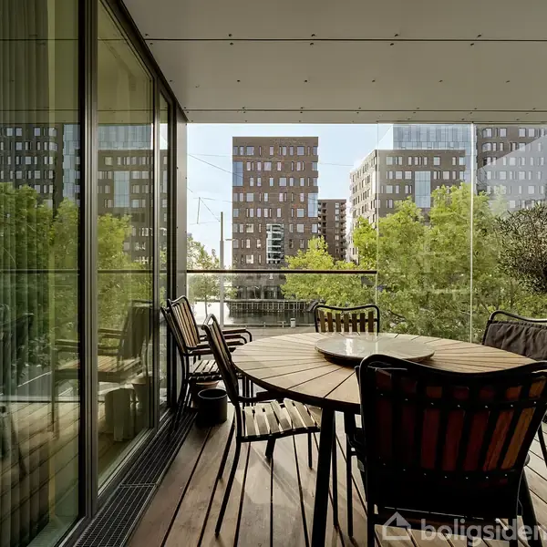 En træterrasse med rundt bord og stole, omgivet af glasvægge. Terrasseplanter står i forgrunden, og moderne bygninger med grøn beplantning ses udendørs i bymiljøet.