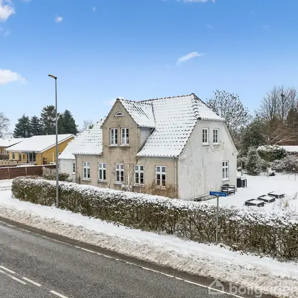 En hvidmalet villa står dækket af sne ved en vej. Sneklædte buske omgiver huset, og flere huse ses i baggrunden under en blå himmel med få skyer.