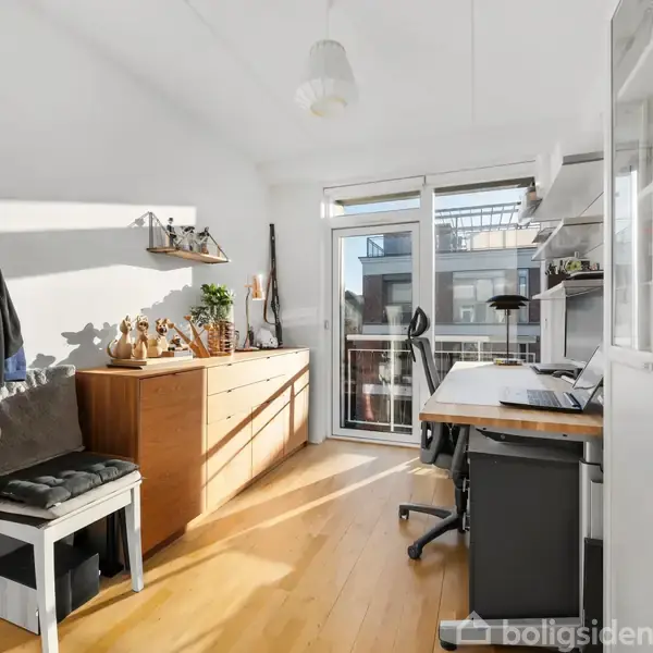 Et kontor har et skrivebord med stol ved et vindue, en jakkestativ med tøj, og en kommode fyldt med dekorative genstande. Værelset er lyst og solrigt.