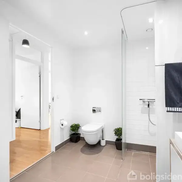 Toilet i moderne badeværelse, monteret på væggen, omgivet af hvide vægge og brunligt gulv. To små planter står på hver side. Til højre ses en bruser og vask med håndklæde.