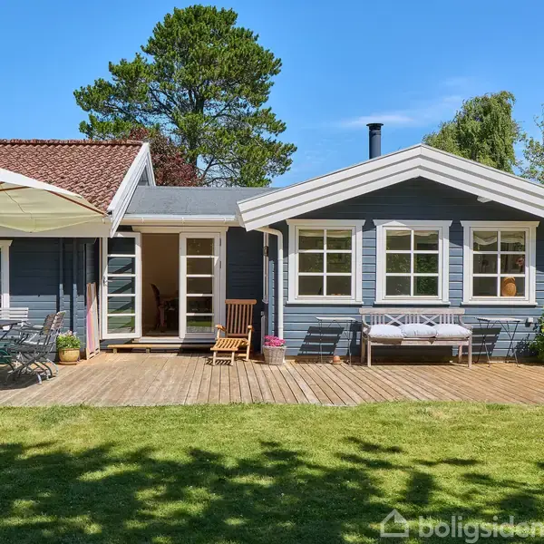 Et blåmalet hus med hvide vinduer står i en grøn have. Der er en træterrasse med havemøbler og en parasol i baggrunden.