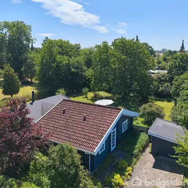 Hus med rødt tegltag omgivet af frodige træer og grøn have. Den blå træfacade har hvide vinduer. Ved siden af huset er en mindre garage. Udsigten viser mange træer i baggrunden.