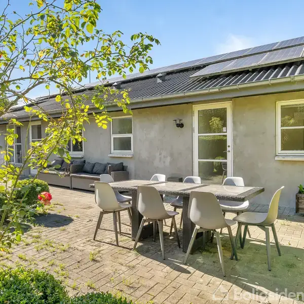 Gråt hus med solceller på taget, udendørs bord og stole placeret på flisebelagt terrasse, omgivet af grønne planter og buske.