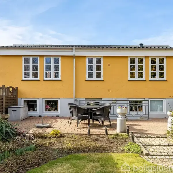 Gul murstensbygning med fire vinduer, placeret i en baghave. En terrasse med havemøbler og brosten omgiver huset. Græsplæne og planter i forgrunden, under en blå himmel.