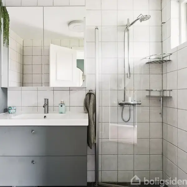 Badeværelse med håndvaskskab i gråt, spejlskab ovenfor, og bruseområde med glasvæg til højre. Hvide fliser på væggene, en plante hænger fra skabet, og et vindue til højre.