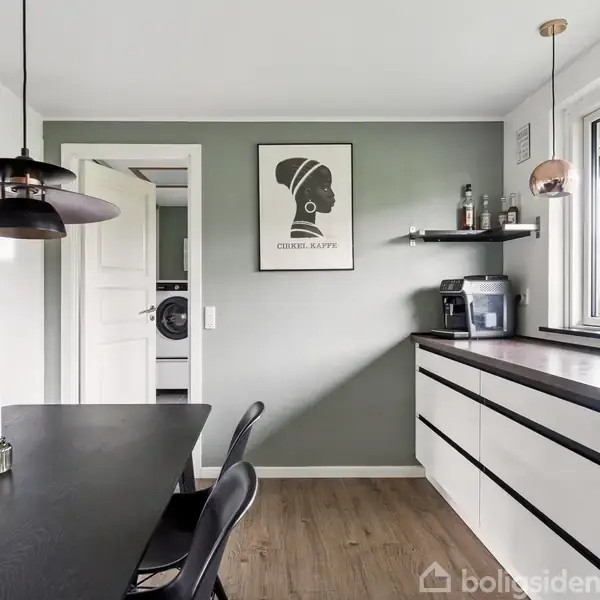 Køkkenbord med sort overflade og stole står under en sort lampe; på væggen hænger en plakat med teksten "CHEEKI KAFFE". I baggrunden ses et køkkenbord med kaffemaskine.