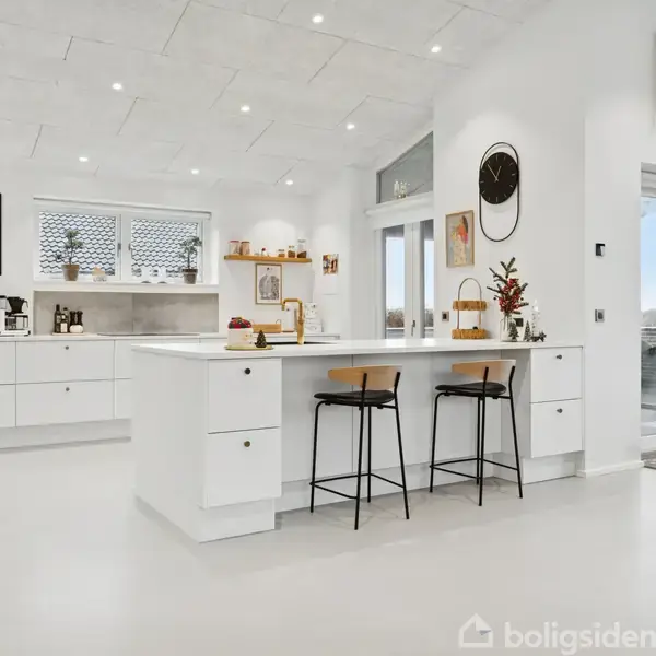 Hvidt køkken med stor ø-bord, over barstole og lyse træhylder. Dekorationer og ur på væggen. Store vinduer og døre giver udsigt til udendørsområde. Kaffe- og køkkenting på bordet.