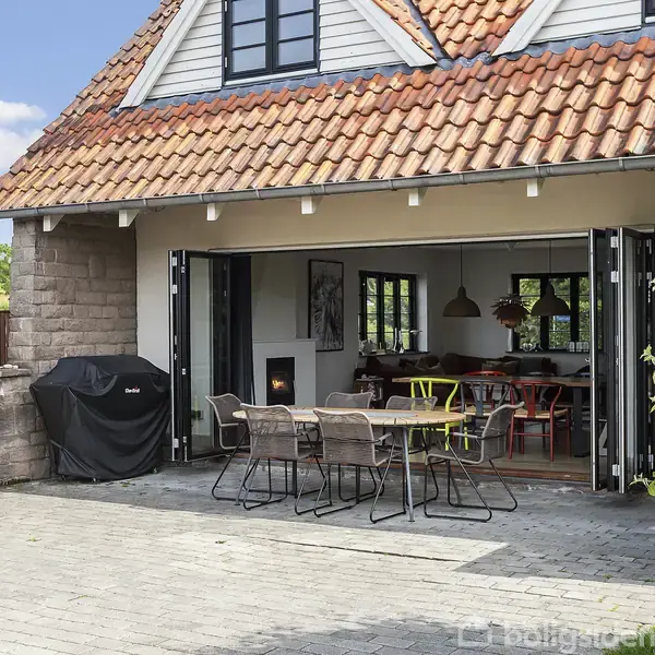 Et udendørs spisebord med seks stole står på en flisebelagt terrasse, grill ved siden af. Åben skydedør fører til hjemmets spisestue, omgivet af grønne planter og have.