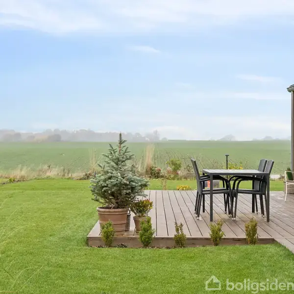 Terrassebord og stole står på et trædæk ved siden af et moderne hus. Omgivet af en velplejet græsplæne med udsigt over grønne marker under en blå himmel.