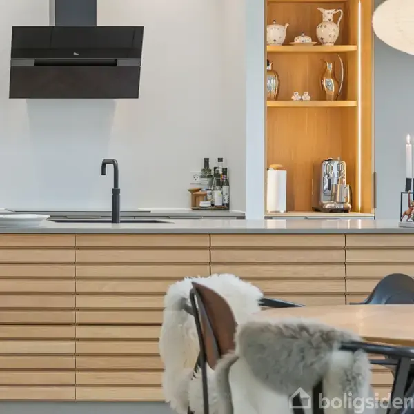 Et moderne køkken med træbordplader og sorte elementer. Et sort emfang ses over en vask. Der er hylder med porcelæn og en kandelaber med tændte lys.