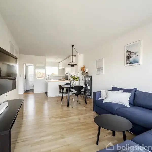 Fladskærmsfjernsyn hænger på væggen, vendt mod en blå sofa i en elegant stue med lyst træ. Spisebord med blomster ses i baggrunden, nær et køkken. Væggene har kunsttryk.