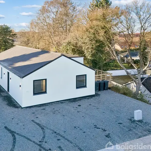 En enkel, hvid bygning med fladt tag ligger på en grusbelagt grund. Bygningen er omgivet af træer og andre huse i et fredeligt, skovklædt nabolag.