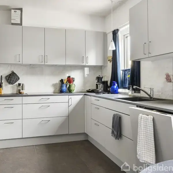 Et moderne køkken med hvide skabe og skuffer. En elkedel og knivblok står på bordet ved siden af en farverig vase. Baggrunden viser vinduer med blå gardiner.