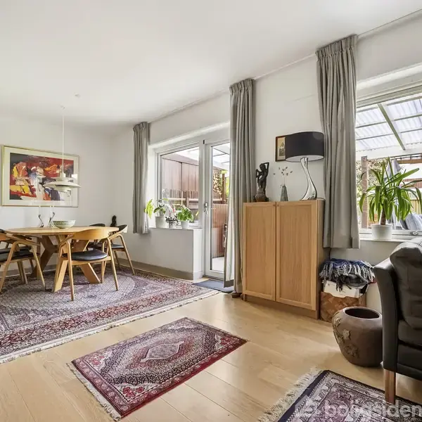 Et lyst stueområde med en spisebordssætning på et orientalsk tæppe. Store vinduer med gardiner slipper lys ind. Planter og dekorative elementer pryder rummet.