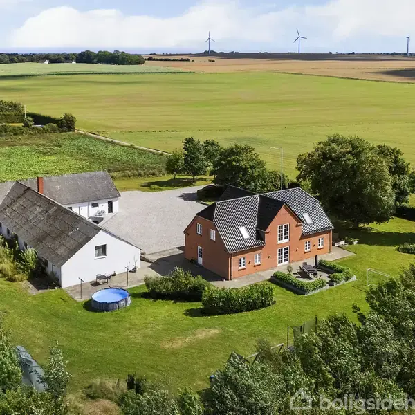 Et rødt murstenshus står ved siden af et hvidt hus omringet af grønne marker og træer. Et rundt, blåt bassin ses på græsplænen nær bygningerne.