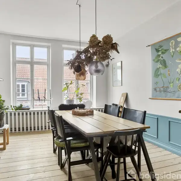 Et langt træbord står i midten af en spisestue omgivet af sorte stole. Over bordet hænger en lampe med tørrede blomster. Rummet har vinduer, grønne planter og en botanisk plakat.