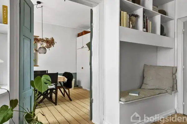 En grøn dør står åben, indrammer udsigt til spisestue med træbord og stole. Til højre er indbygget bænk med puder, bøger på hylde ovenover. Plante dekorerer rummet.