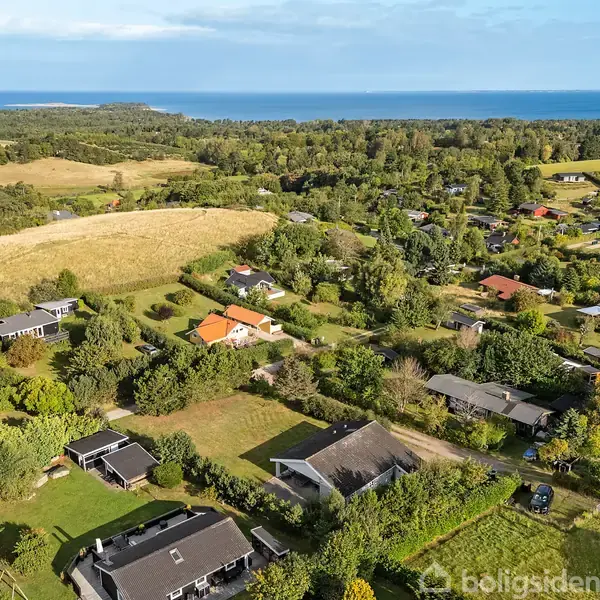 Landsbyhuse spredt over grønne marker og skov, med kystlinje og hav i baggrunden, hvilket skaber et fredeligt, landligt miljø.