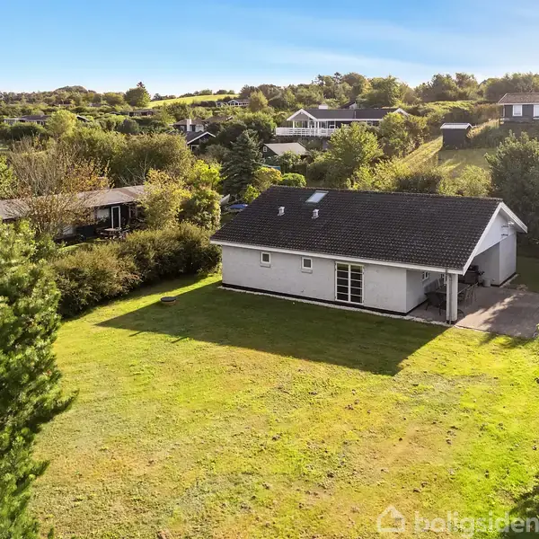 Et hvidt hus med sort tag står på en stor, grøn græsplæne omgivet af træer og andre huse i et bakket landskab under en blå himmel.