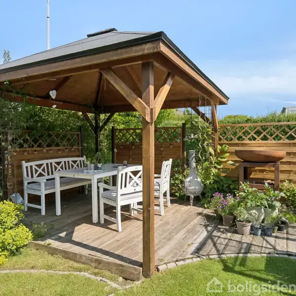 En åben pavillon i træ med et hvidt bord og bænke. Omkring den er en velplejet have med planter og et drivhus i baggrunden under en blå himmel.