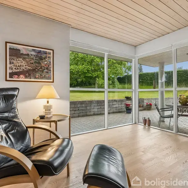 En carport med et tegltag står ved siden af et murstenshus. Omgivelserne består af en grøn have med træer og en hæk.