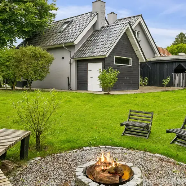 Bål i en rund bålplads med brændende flammer, omgivet af grus og to træstole. Græsplæne og træer omkranser et moderne hus med grå tagsten og hvid facade.