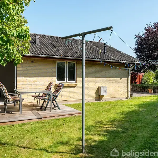 Terrassen ved huset har havemøbler, mens tøjsnoren står i græsplænen omgivet af grønne buske og træer. Himlen er klar, hvilket skaber en lys og indbydende atmosfære.