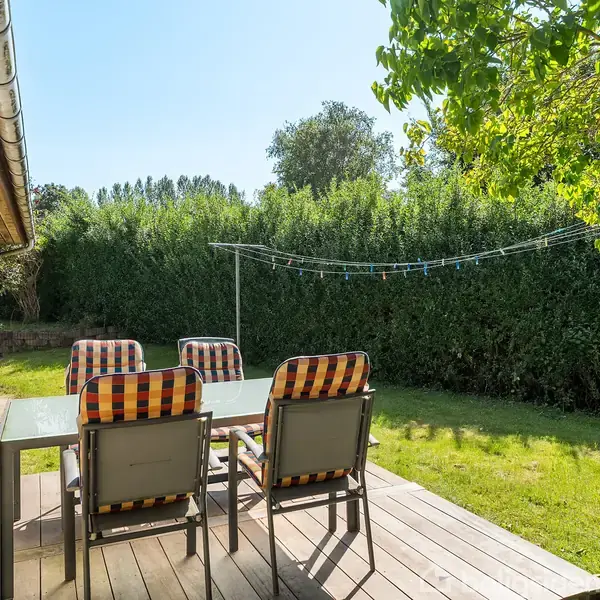 En terrasse med et bord og fire stole står med stribede hynder. Den ligger ved et hus, omgivet af en græsplæne og tætklippede buske.