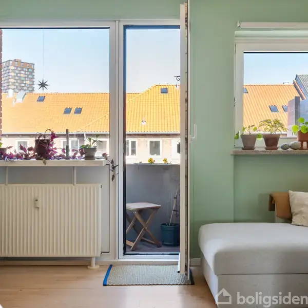 Stue med en hvid sofa til højre og en balkon dør i midten, planter på vindueskarmen, grønne vægge og udsigt til orange tagbygninger udenfor. Ingen tekst findes i billedet.