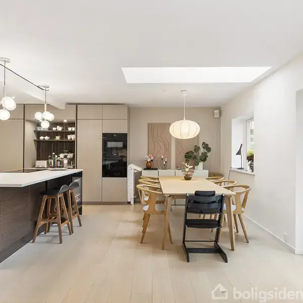 Køkken-alrum med stort spisebord og stole, moderne køkkenø med barstole. Belysning med runde lamper og ovenlys. Lyse gulve og store vinduer giver masser af naturligt lys.