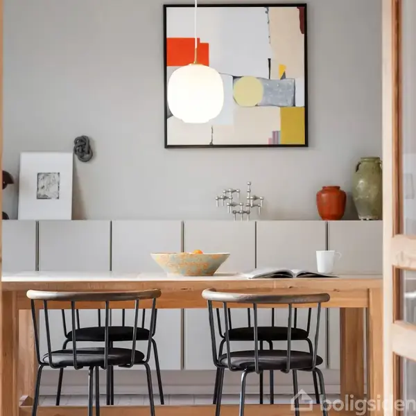 En træspisebord med to sorte stole står i en moderne spisestue. En skål og en kop er på bordet. En abstrakt maleri hænger på væggen, og en lampe lyser over bordet.