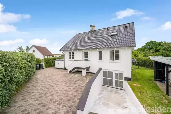 Et hvidt hus med mørkt tag står stille omgivet af en belagt gård og grønne hække. Græsplæne og træer ses i baggrunden under en blå himmel med få skyer.