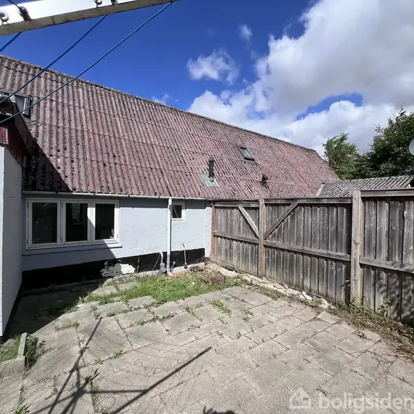 En brolagt gårdsplads med en træport og et hvidt hus med rødt tegltag, omgivet af et plankeværk. Blå himmel og hvide skyer ses i baggrunden.