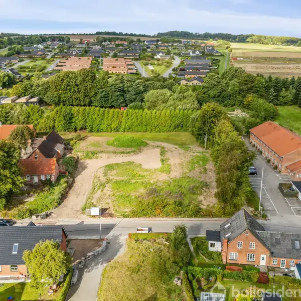 En byggegrund ligger midt i en landsby omgivet af huse med røde tage og grønne marker i baggrunden. Der er træer omkring og små veje leder gennem området.