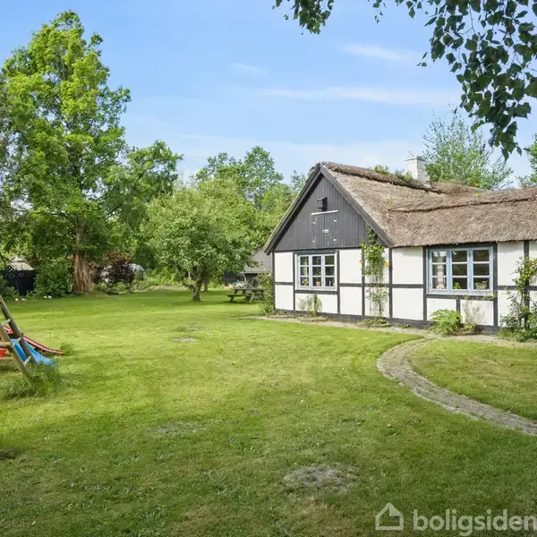 Det bindingsværkshus med stråtag står stille i en grøn have, flankeret af store træer. Ved siden af er en gyngestativ med rutsjebane, der skaber en hyggelig atmosfære.