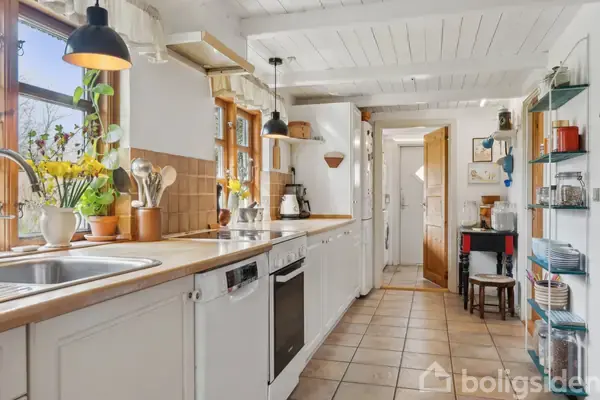 Køkkenbord med hvidt kabinet og køkkenvask, omgivet af træskærebræt og køkkenredskaber. Lyse pendel-lamper over bord, store vinduer med planter, og en åben trædør i baggrunden.