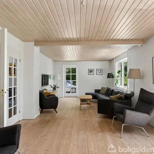 Stue med moderne møbler, inklusive en grå sofa og to stole, der står på et trægulv. Hvide vægge og loft med indbyggede spots. Store vinduer giver udsigt til en have.