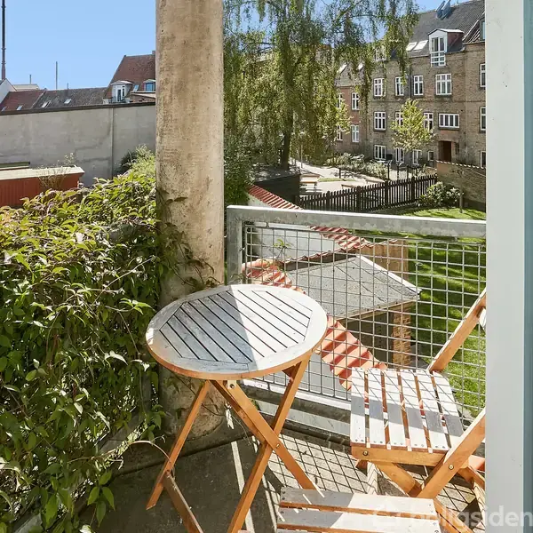 Rundt havebord står på altan med klapstol ved siden af. Altanen har trådhegn og udsigt til en gårdhave med græs og bygninger i baggrunden.